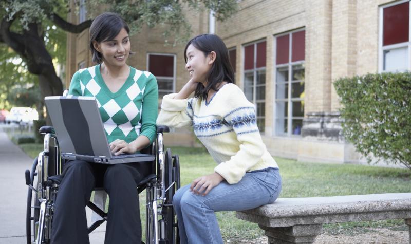 One female student sitting on a concrete bench studying with another female student in a wheelchair holding a laptop on her lap